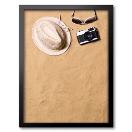 Obraz w ramie Okulary przeciwsłoneczne, wiklinowy kapelusz i aparat fotograficzny na piasku