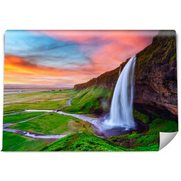 Fototapeta Piękny krajobraz z zielonymi równinami i wysokim wodospadem wylewającym się z pokrytej mchem kamiennej ściany